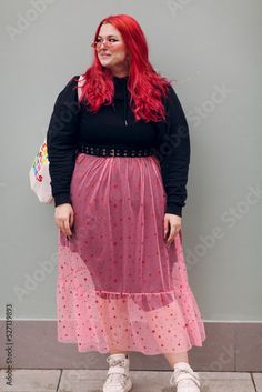 a woman with red hair and glasses is standing in front of a gray wall wearing a pink polka dot skirt