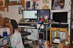 a cluttered room with two computer monitors and many other items on the desks