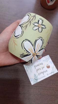 a hand holding a cup that has been decorated with flowers and leaves on it, next to a business card