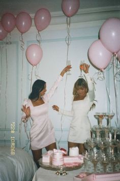 two women in white dresses holding pink balloons and standing next to each other with cake on the table