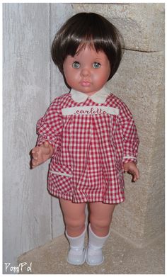 the doll is wearing a red checkered dress and white socks with her hands in her pockets