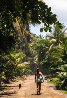 a woman walking down a dirt road with a surfboard in her hand and a dog on the other side