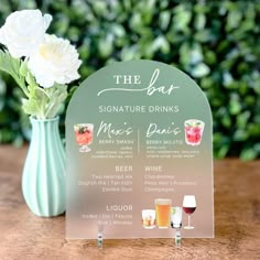 a sign with drinks on it sitting next to a vase filled with flowers and greenery