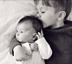 two young children cuddle together on a white sheet, one holding the baby's head