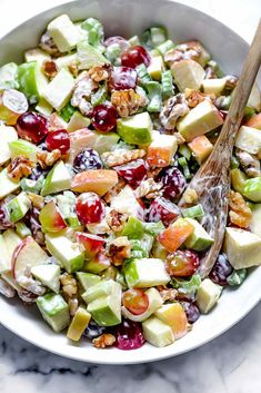 a white bowl filled with apples, grapes and walnuts next to a wooden spoon