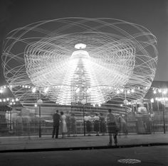 people are standing around in front of a ferris wheel at night with lights on it