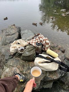 a person holding a cup of coffee on top of a rock next to a body of water