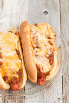 two hot dogs covered in cheese and chili sitting on a glass plate next to each other