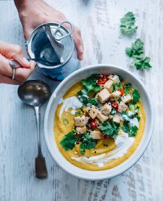 two hands holding a bowl of soup with tofu and cilantro on the side