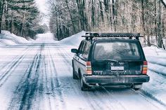 a car driving down a snow covered road in the middle of winter with trees on both sides