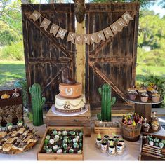 a table topped with cakes and desserts next to a wooden fence covered in cactus