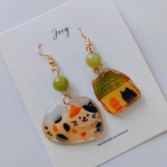 two earrings with cats on them sitting next to each other, one has a green bead