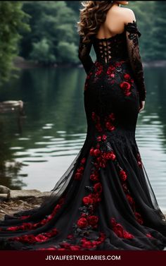 a woman in a long black dress with red flowers on the skirt is standing near water