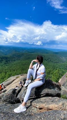 a woman sitting on top of a large rock next to a lush green forest under a blue sky