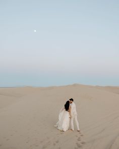 a bride and groom standing in the sand dunes at their desert wedding venue, with moon rising above them