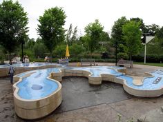 an outdoor swimming pool with benches and people in the background, surrounded by green trees