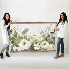 two women standing next to each other in front of a floral wall hanging on the wall