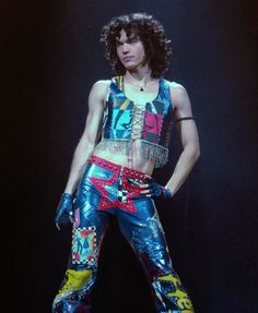 a man with long curly hair wearing colorful pants and holding his hands on his hips