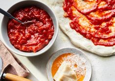 two pizzas with sauce in bowls and one on the table next to it is an uncooked pizza