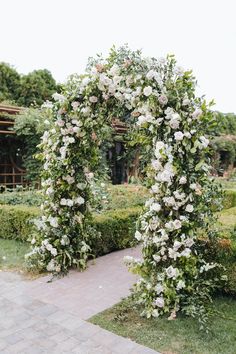 an arch covered in white flowers next to a brick walkway and garden area with hedges