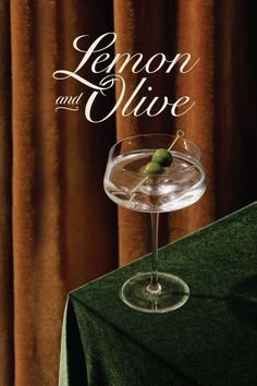 lemon and olive in a martini glass on a green table cloth with curtains behind it