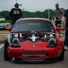 two men standing next to a red sports car with the hood up and engine on