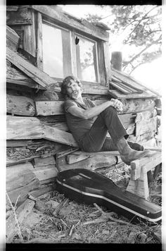 black and white photograph of a man sitting on a bench in front of a log cabin