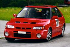 a red car driving down a street next to tall grass and trees in the background