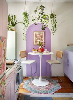 a small kitchen with purple walls and white table in the center, plants hanging from the ceiling