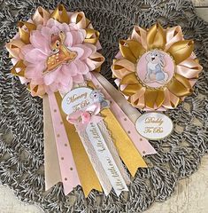 two pink and gold ribbon flowers sitting on top of a metal tray with tags attached to them