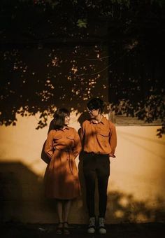 two people standing next to each other in front of a tree and wall with shadows on it