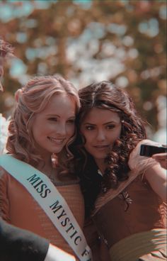 two women standing next to each other in front of some trees and one is holding a camera