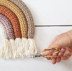 a person holding scissors near a crocheted rainbow wall hanging on a white wall