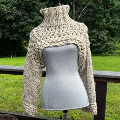 a mannequin wearing a knitted sweater in front of a wooden fence with trees and grass behind it