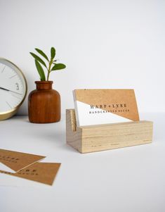 a wooden business card holder next to a small plant and clock on a white surface