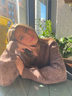 a woman sitting at a table with her hand on her chin and looking into the camera