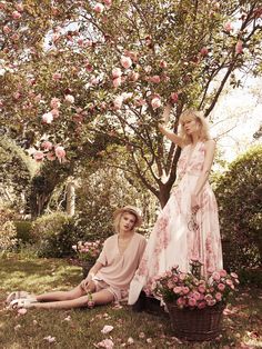 two women sitting on the ground in front of a tree with pink flowers around them