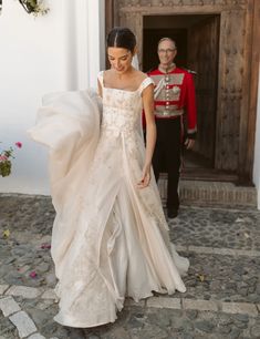 a woman in a wedding dress walking down the street with a man in uniform behind her