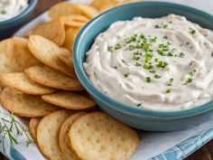 a blue bowl filled with white dip surrounded by crackers on a napkin next to it