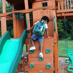 a young boy climbing up the side of a wooden play structure with green slide and slides