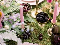 the table is set with candles, flowers and fruit in bowls on mossy ground