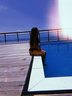 a woman sitting on the edge of a swimming pool looking out at the ocean with her back to the camera