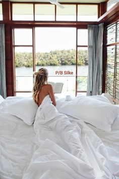 a woman sitting on top of a bed covered in white sheets and pillows looking at a laptop computer