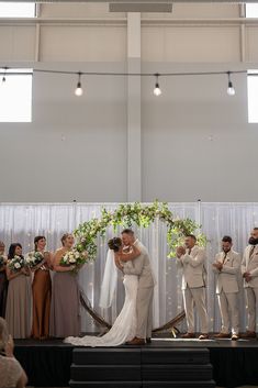 a bride and groom kissing in front of their wedding party on the stage at an indoor ceremony