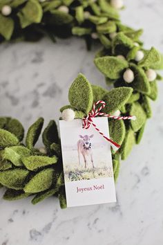 a christmas wreath with a card attached to it on a marble surface, decorated with green leaves and white balls