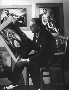 black and white photograph of man sitting in chair with paintings on the wall behind him