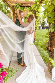 a bride and groom standing under a pergolated arbor with their veil blowing in the wind