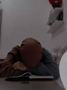 a person wearing a brown hoodie is sleeping on a desk with a book in front of them