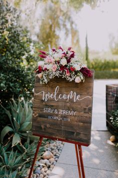 a welcome sign with flowers and greenery on it in front of some bushes at a wedding