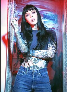 a woman with long black hair and tattoos standing in front of a mirror holding an object